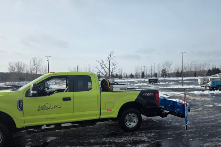 A lime green Shelton maintenance truck with a blue salt spreader attached is parked in a snowy lot. Leafless trees and utility vehicles are visible in the background.
