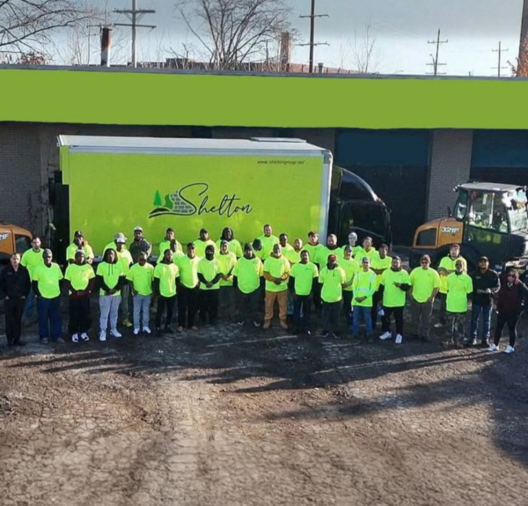 A group of people wearing neon yellow shirts stand in front of a green and yellow truck and construction equipment on a dirt lot.
