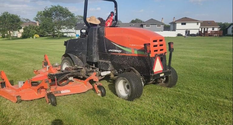 A large red and black lawn mower with an orange deck sits on a grass field near residential houses, its seat adorned with a straw hat—a perfect sight for those seeking reliable sod service near me.