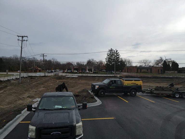 A construction site features a parked black truck and trailer in the foreground, with power lines and trees silhouetted under an overcast sky. Nearby, Homewood sod installation services are transforming landscapes, perfect for completing projects with aesthetic grassy expanses.
